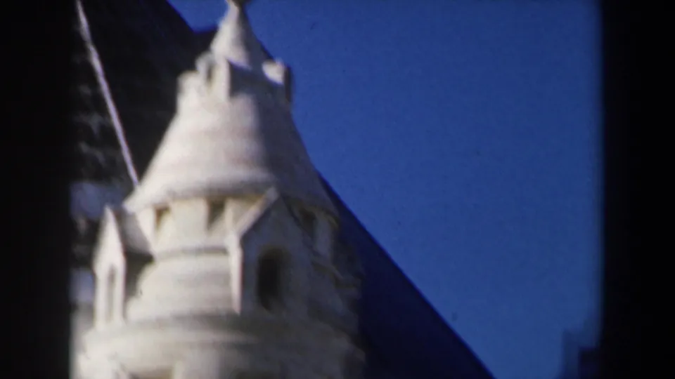 1960 Israel 8mm frame with a pale church spire, arched openings, and adjacent weathered roof edge against blue sky.