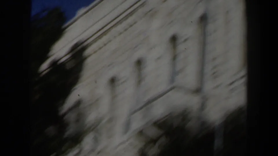 Handheld 1960 Israel frame with motion blur across a white stone building, arched windows, and dark foreground shadows.
