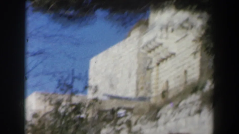 1960 Israel 8mm frame showing a sunlit white stone building corner, arched windows, blue sky, and tree branches.