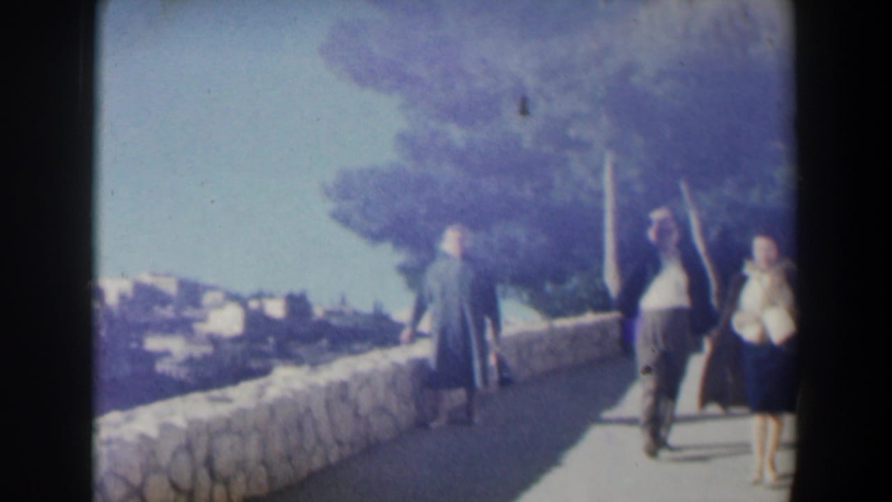 1960 Israel home-movie frame showing adults on a sunlit path near stone walls, trees, and distant hillside buildings.