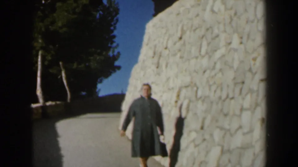 1960 Israel footage frame showing a woman in a grey coat walking along a curved path beside a tall white stone wall.