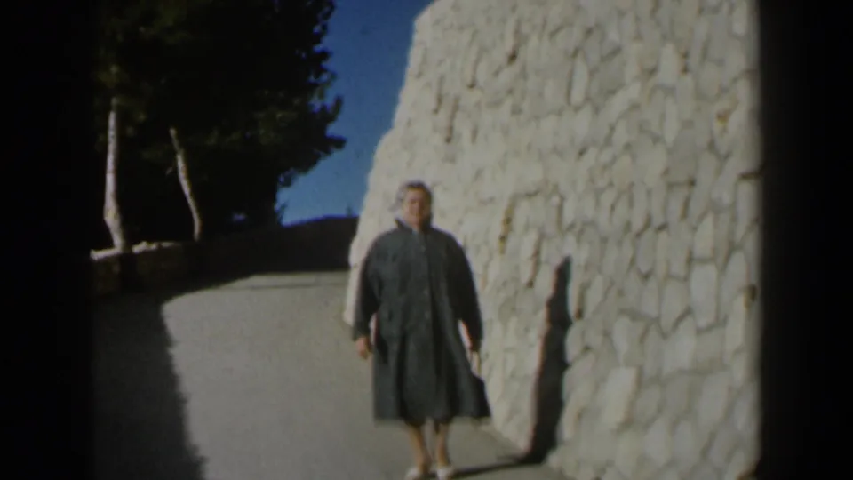 The woman in a grey coat is framed closer beside the white stone wall, with trees and blue sky in the background.