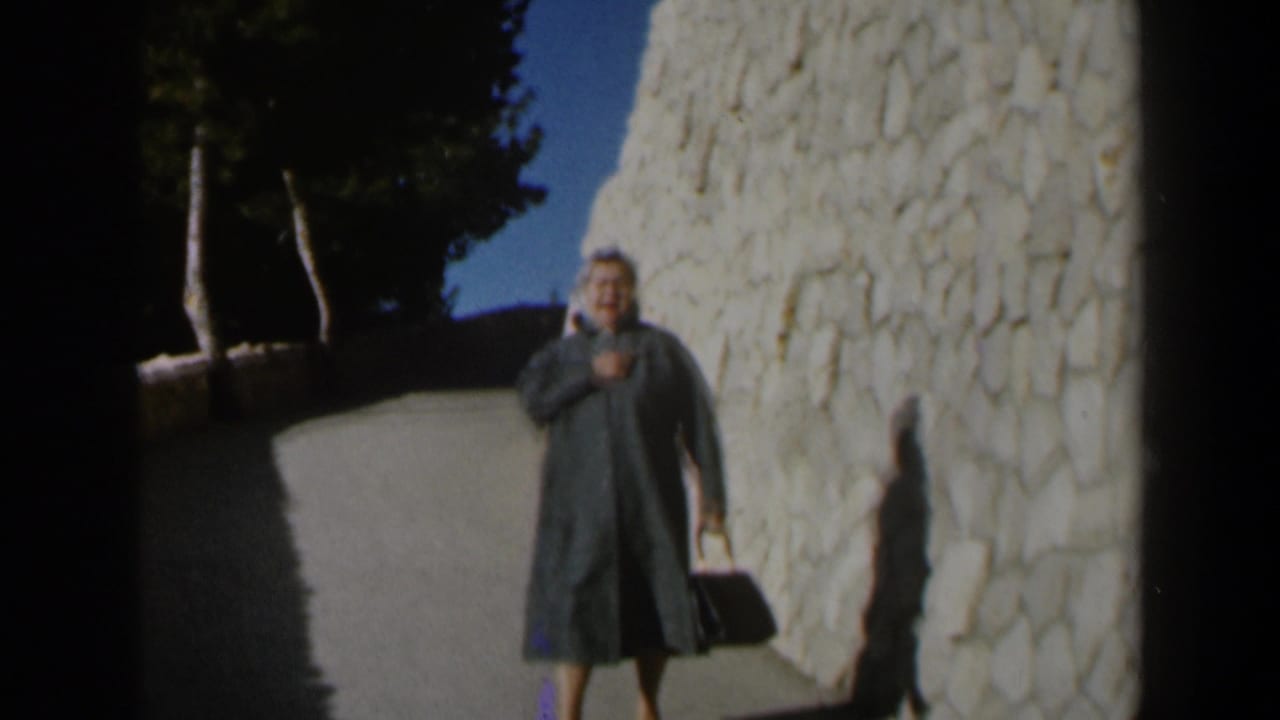 In 1960 Israel, a woman in a dark coat walks toward the camera along a bright stone pathway with trees overhead.