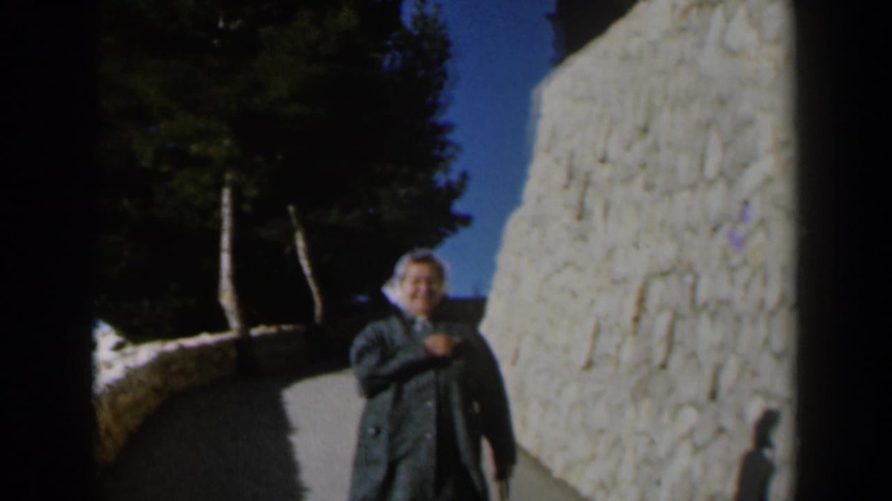 The woman smiles while moving closer along the Israel walkway, framed by a pale stone wall, trees, and blue sky.