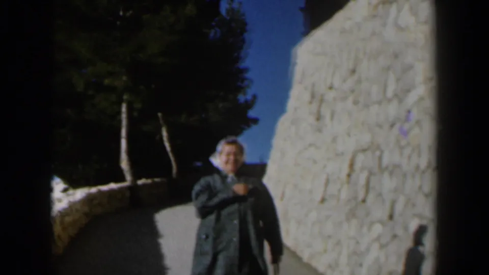 The woman smiles while moving closer along the Israel walkway, framed by a pale stone wall, trees, and blue sky.