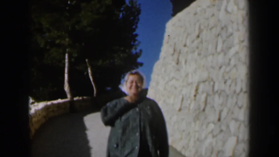 A smiling woman in a dark coat nears the 8mm camera on a sunlit path in Israel, with a textured stone wall behind her.