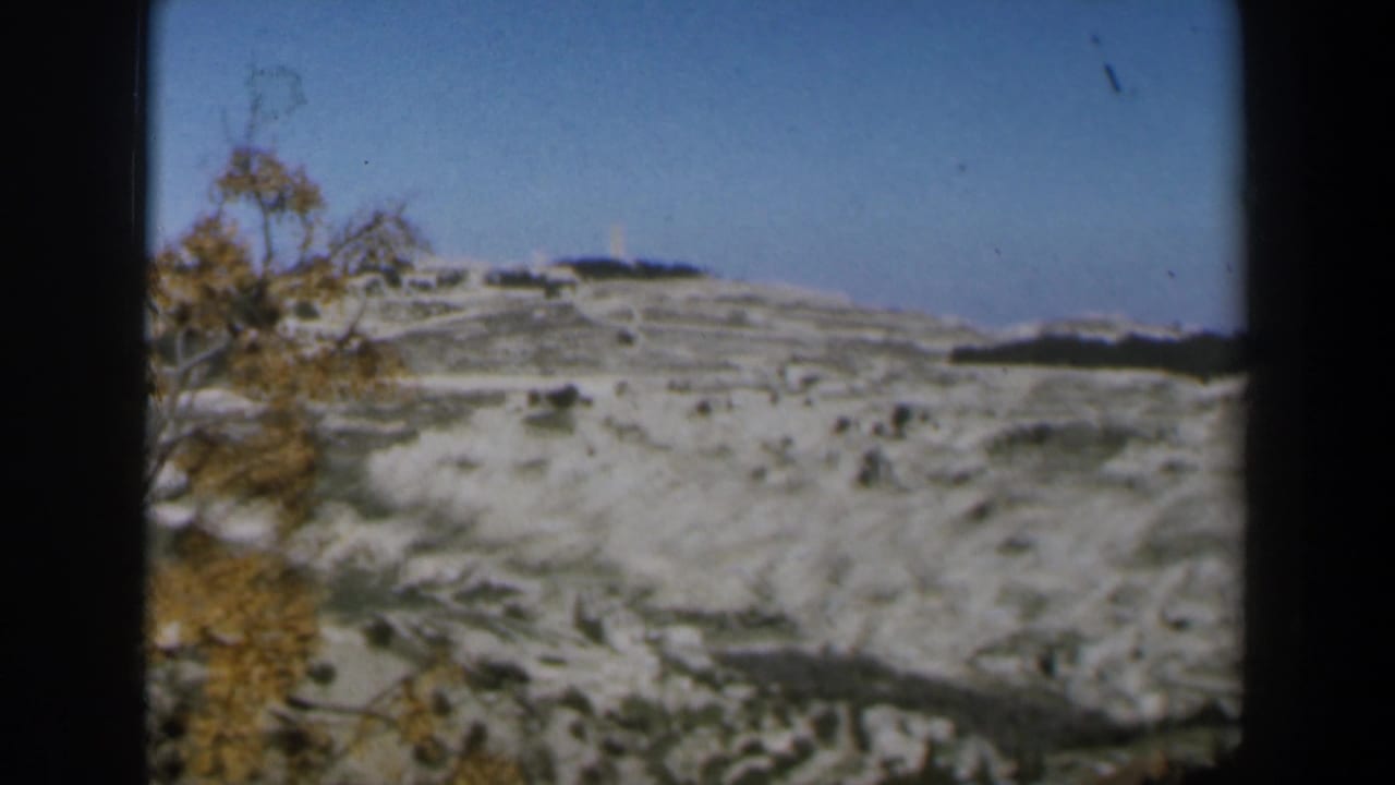 1960 Israel 8mm frame showing a pale, snow-dusted rocky hillside beneath blue sky, with autumn-colored tree branches along the side.