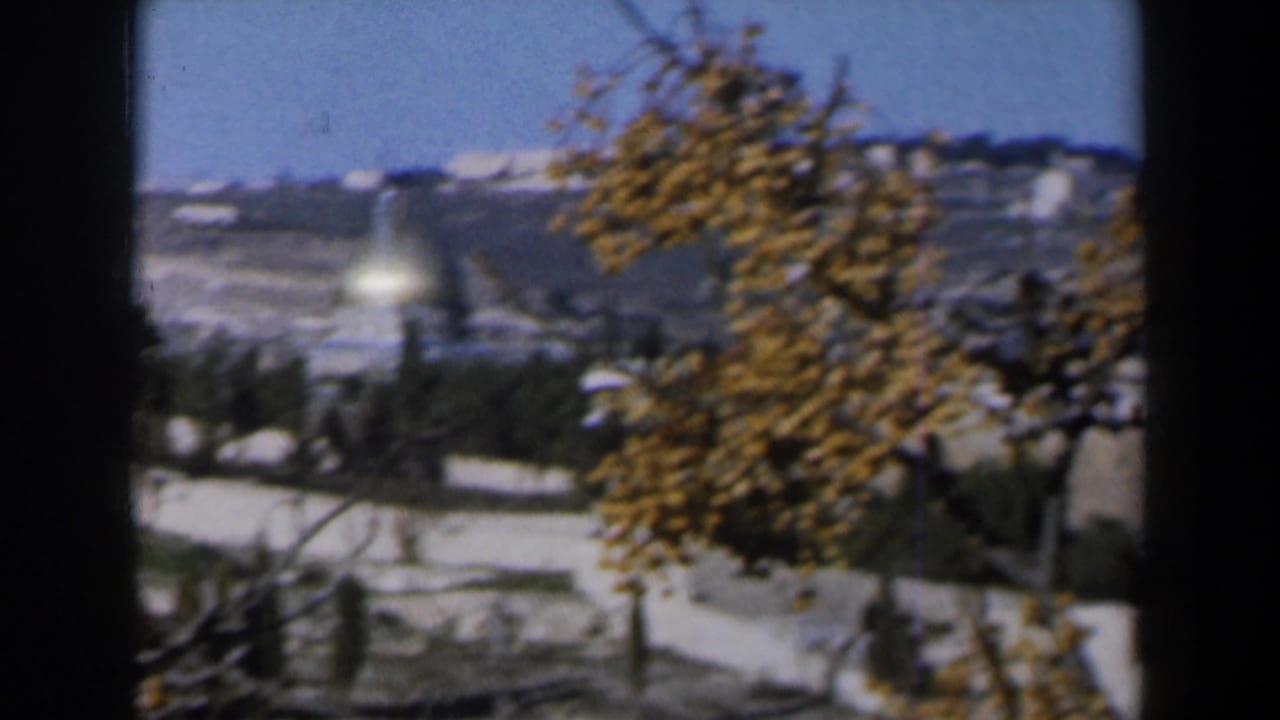 1960 Israel home-movie view across pale walls and autumn trees toward a snow-marked hillside and distant light-toned structures.