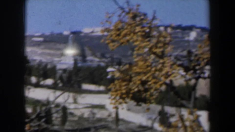 1960 Israel home-movie view across pale walls and autumn trees toward a snow-marked hillside and distant light-toned structures.