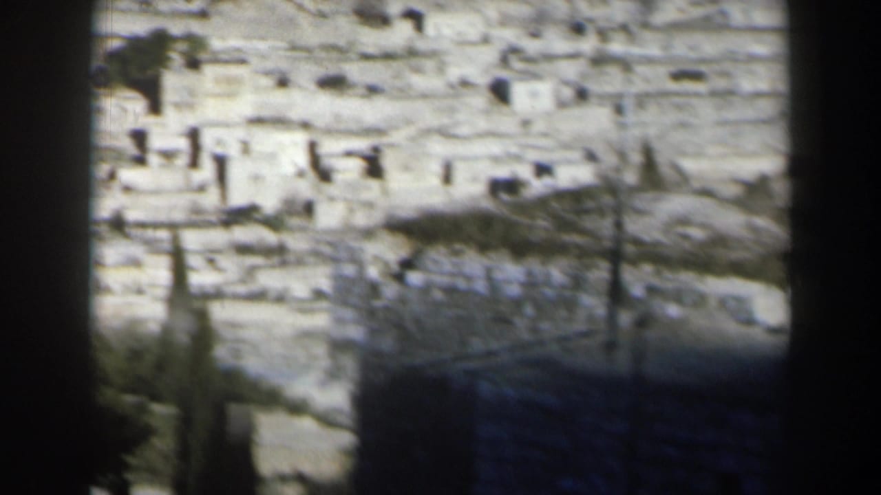 Sunlit Israeli hillside in 1960 with scattered pale buildings, rough stone ground, and a dark vertical frame edge.