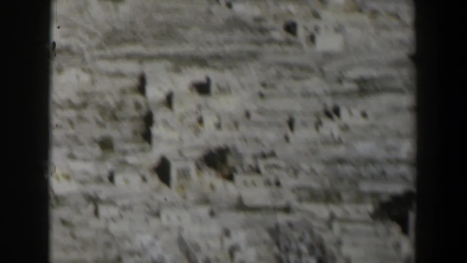 Blurred 8mm frame of an Israeli hillside in 1960, with small pale structures and uneven stone terrain.