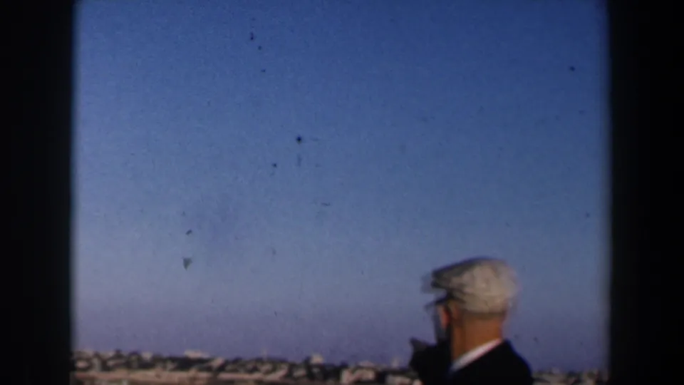 1960s Israel home-movie frame showing a man in a white cap near the bottom edge, facing a distant town under open sky.
