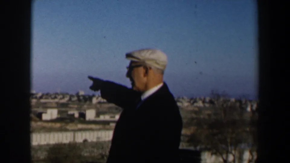 1960s Israel 8mm frame of an adult man in profile pointing across a distant low-rise town under a blue sky.