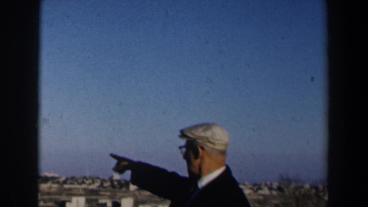 Elderly man in a flat cap points toward a distant town under a blue sky.