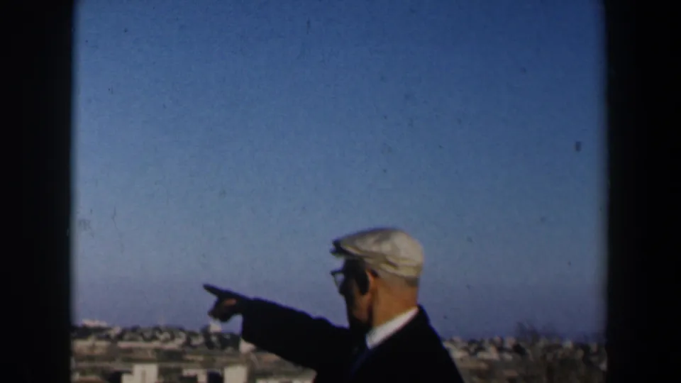 1960 Israel home-movie frame showing an elderly man in glasses and a flat cap pointing from a high viewpoint toward buildings below.