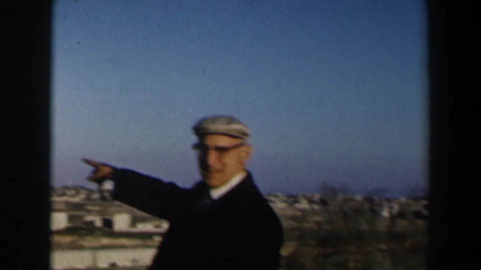 1960 Israel home-movie frame of an elderly man in a dark jacket and flat cap pointing toward a town from a raised viewpoint.