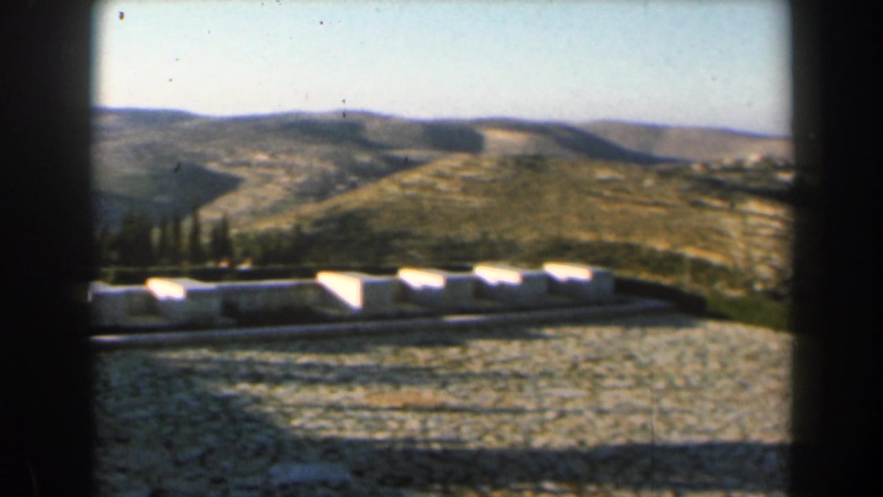Arid hills beyond pale stone ground and low white structures.