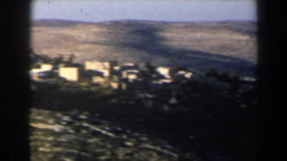 Soft 8mm frame of an Israeli village area below broad hills, with trees and pale buildings partly in shadow.