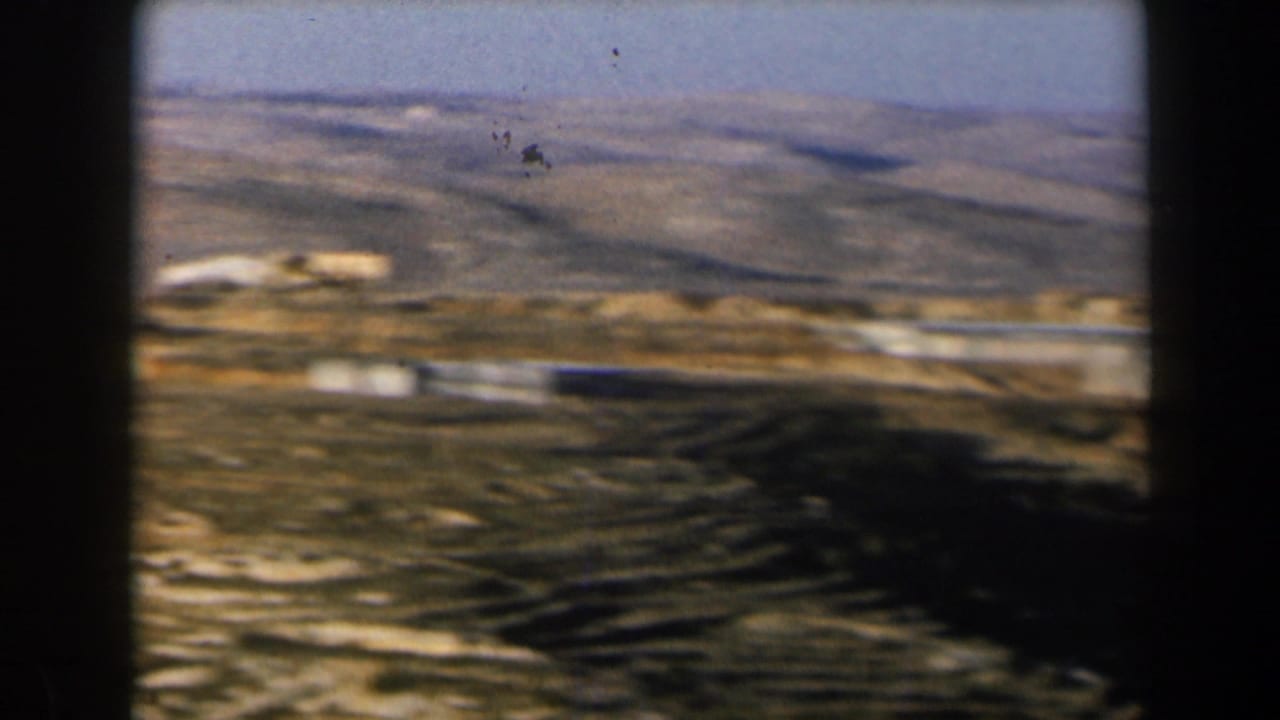 Dry hillside landscape with distant buildings, roads, and dark vertical frame edge.
