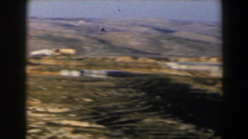 1960 8mm Israel landscape with distant white buildings, winding roads, terraced ground, and arid hills under blue sky.