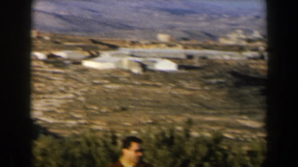 1960 Israel 8mm frame showing low white buildings, dry slopes, scrub vegetation, and a partial foreground figure at the bottom edge.