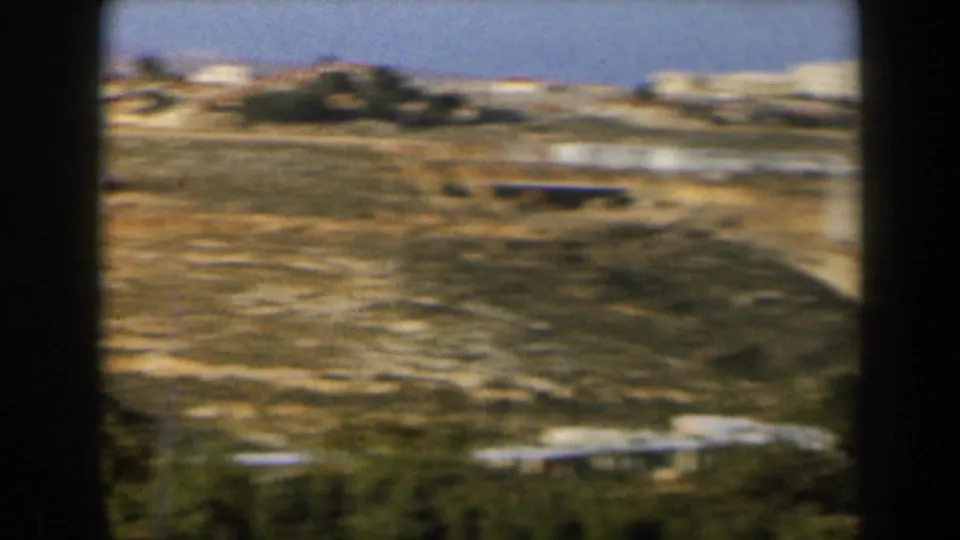 1960 8mm view in Israel of scattered white structures, pale roads, dry ground, and rolling hills in soft faded color.