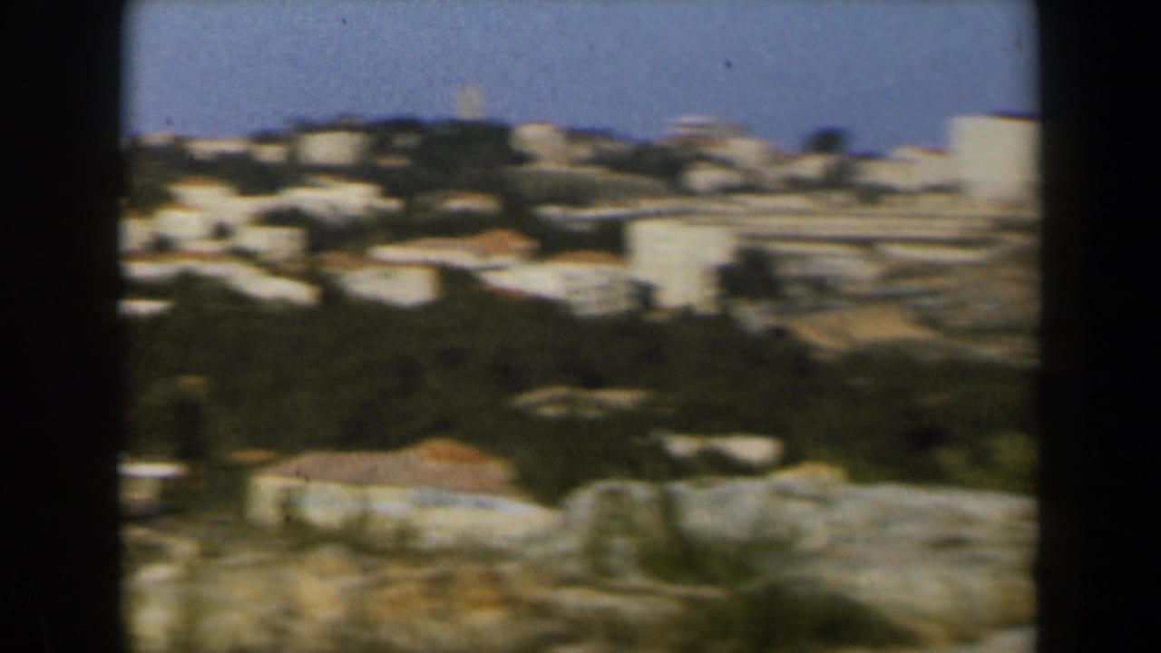 Soft 8mm frame of an Israeli hillside town, with pale buildings and tree cover beneath a clear blue sky.