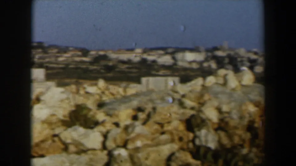Handheld 8mm view of rocks in the foreground and a distant Israeli hillside town under blue sky.