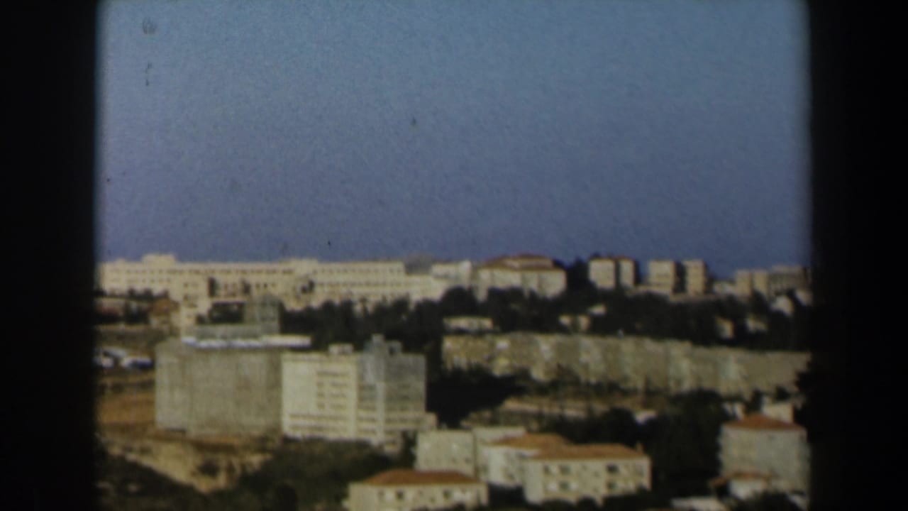 1960 Israel 8mm frame showing an elevated view across pale buildings, tree lines, dry ground, and a clear blue sky.