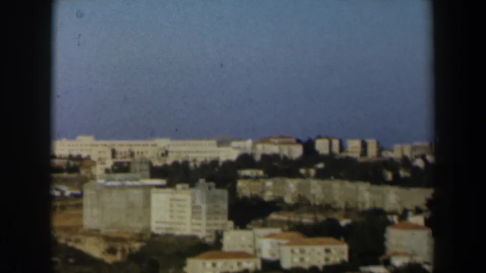 1960 Israel 8mm frame showing an elevated view across pale buildings, tree lines, dry ground, and a clear blue sky.