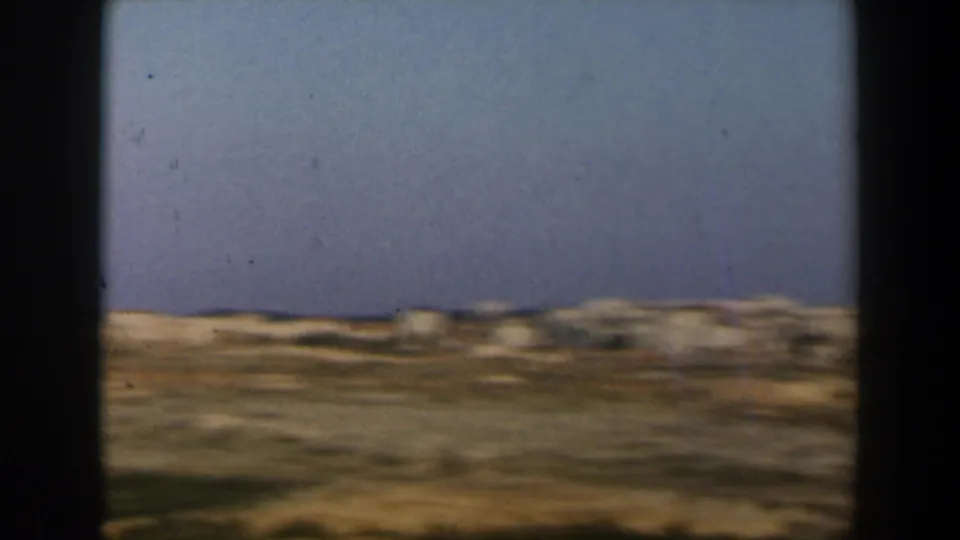 1960 Israel travel-film frame with open dry slopes, distant pale structures, and a wide blue sky during the pan.