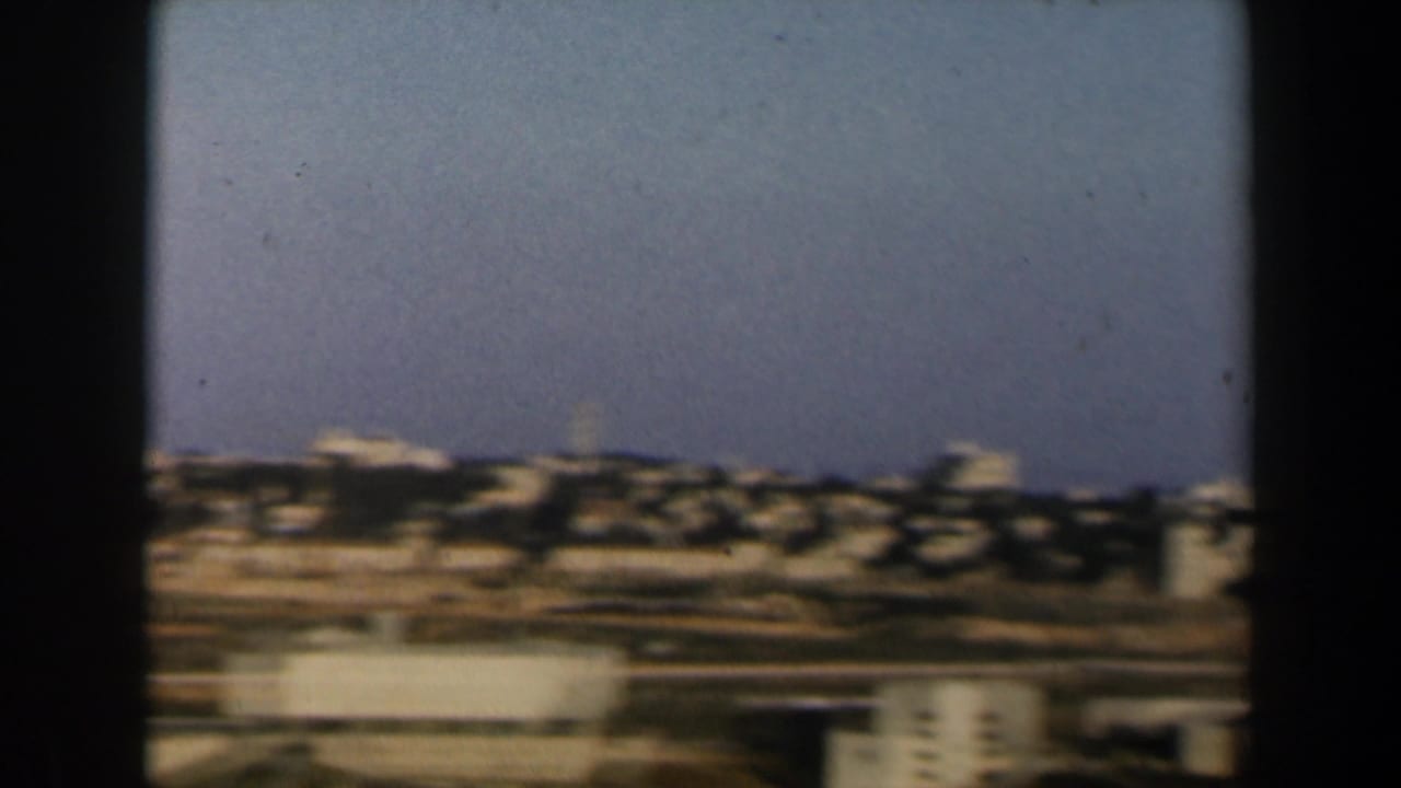1960 Israel home-movie frame showing a blurred hillside townscape with tan buildings, open ground, and blue sky.