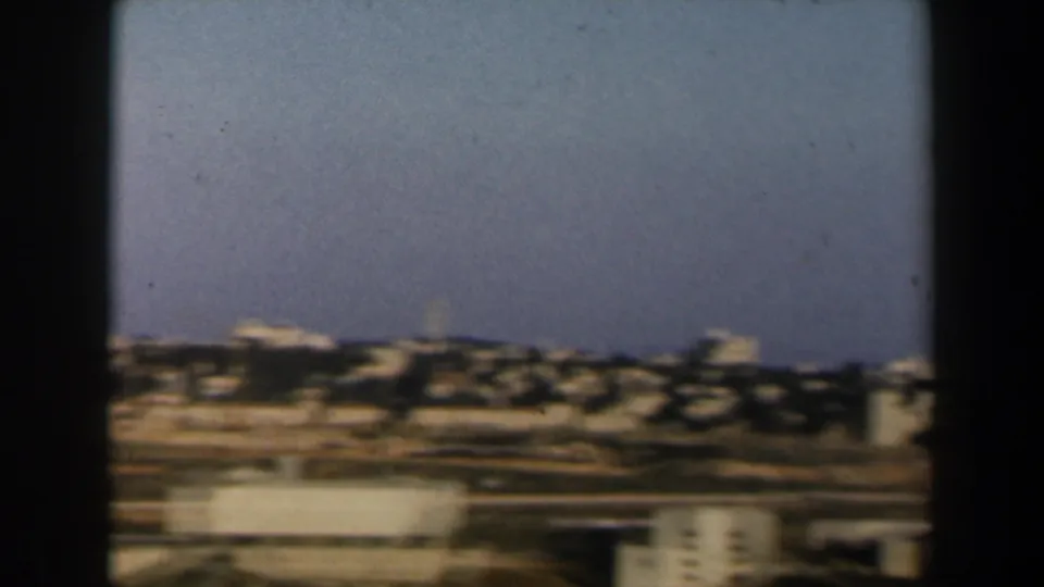 1960 Israel home-movie frame showing a blurred hillside townscape with tan buildings, open ground, and blue sky.