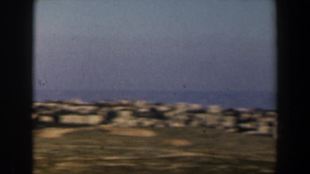 1960 Israel frame with a dense band of pale buildings across low hills, softened by handheld motion and film grain.