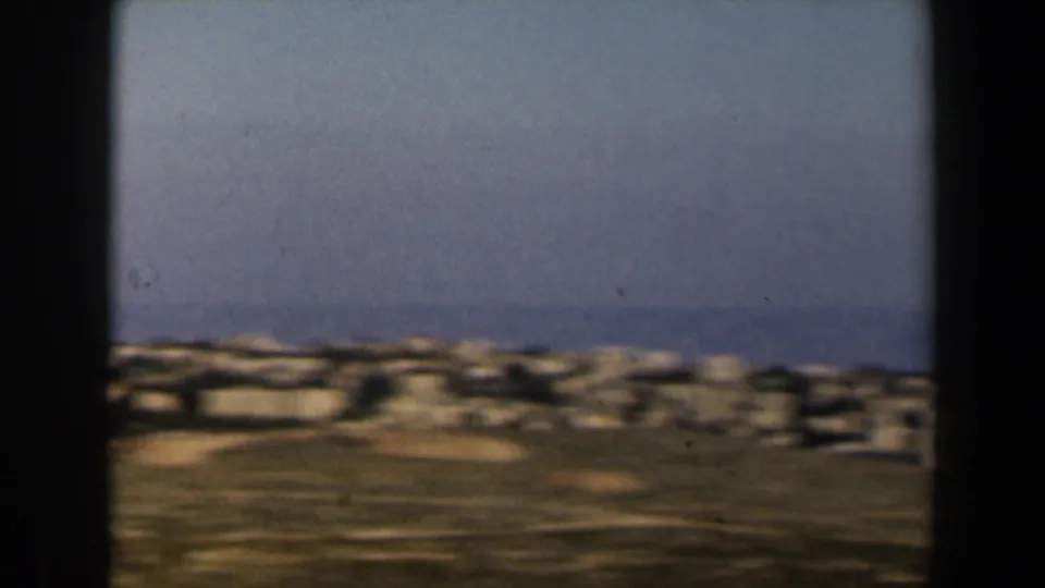 1960 Israel frame with a dense band of pale buildings across low hills, softened by handheld motion and film grain.