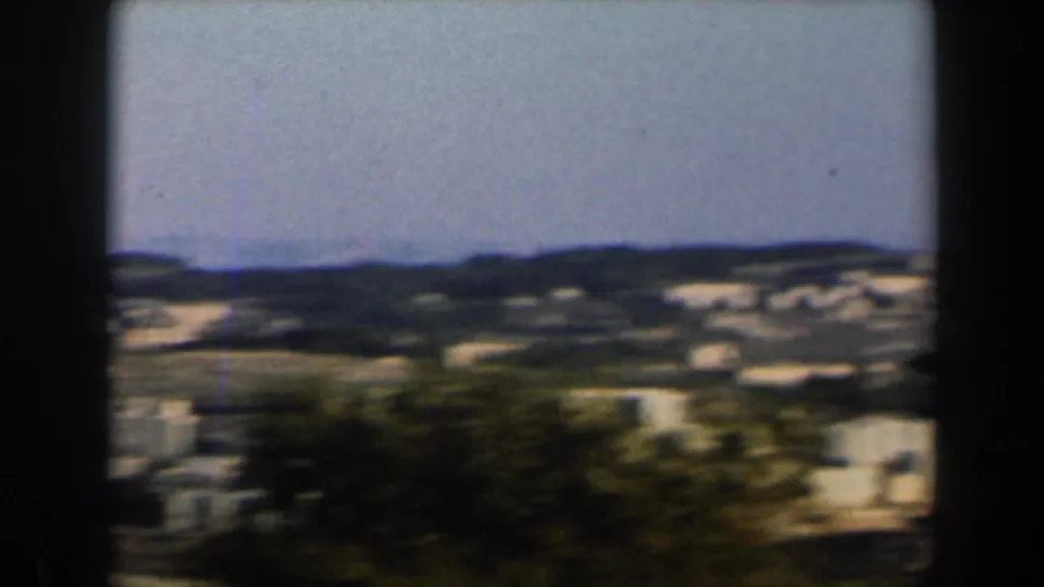 1960 Israel frame showing pale buildings, foreground trees, and green-brown hills beneath a clear sky.