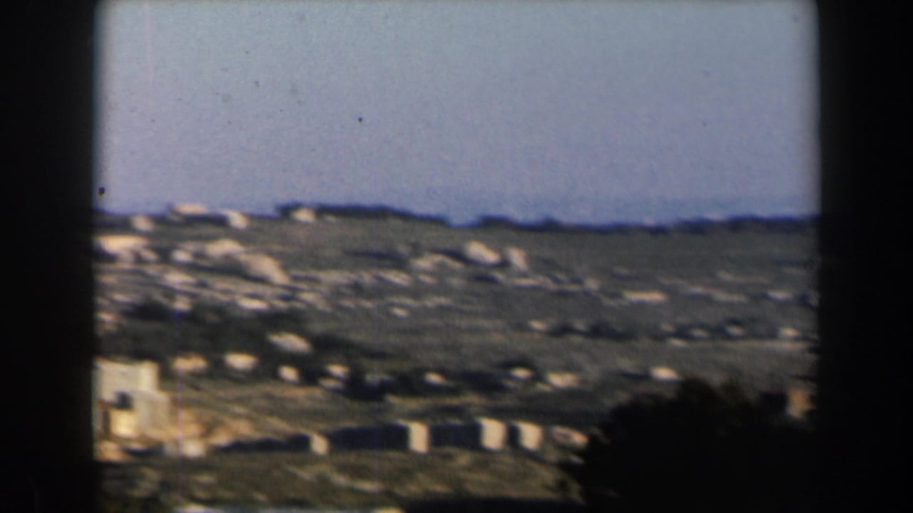 1960 Israel 8mm frame showing a rocky, sunlit hillside with low pale structures beneath a wide sky.