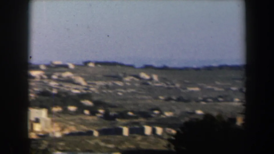 1960 Israel 8mm frame showing a rocky, sunlit hillside with low pale structures beneath a wide sky.