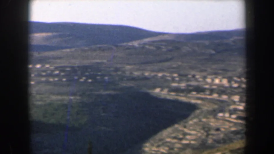 1960 Israel 8mm frame showing a broad valley settlement, pale roads or paths, hills, and dark gate edges.