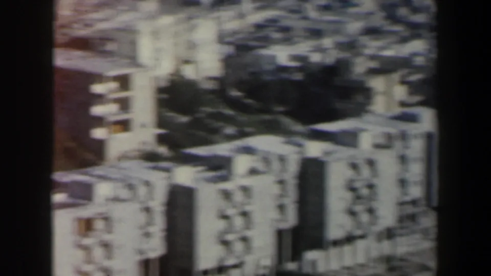 Aerial home-movie view in Israel showing block-style residential buildings, shadowed open ground, and soft 8mm color fading.