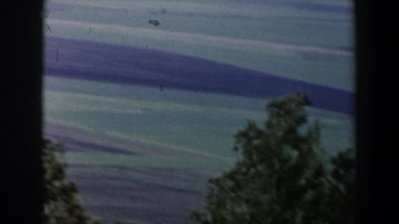 1960 Israel home-movie frame with rolling grassy hills, distant ridges, and dark foreground trees under a muted sky.