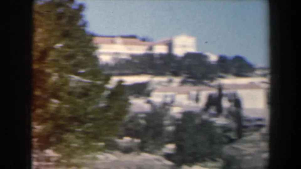 1960 Israel 8mm view of a hillside building complex seen through trees near a lake or mountain landscape.