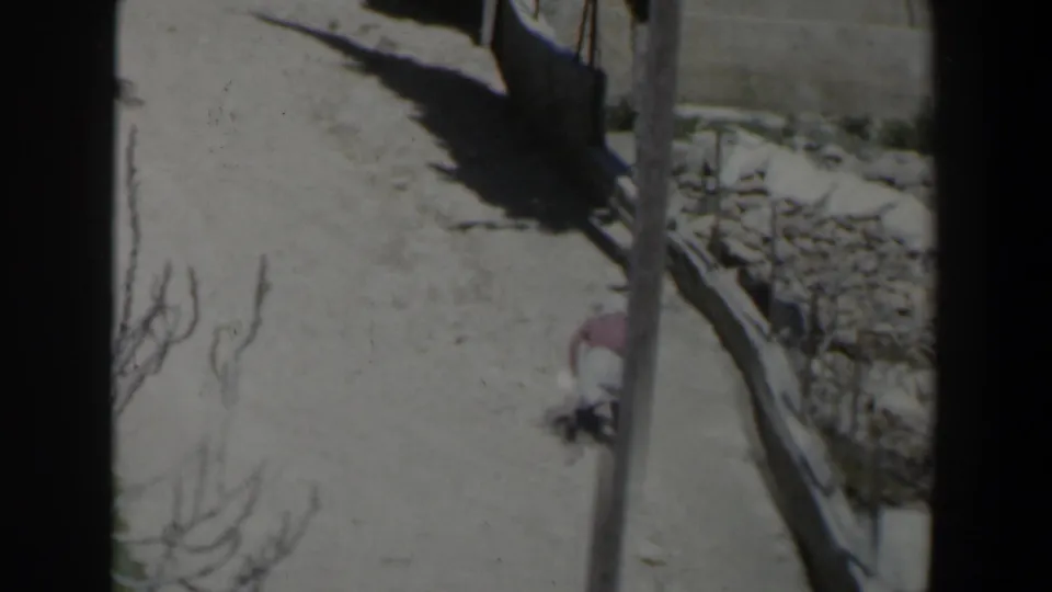 1960 Israel 8mm frame showing a veiled woman moving along a sunlit path beside rough stone walls and dry ground.