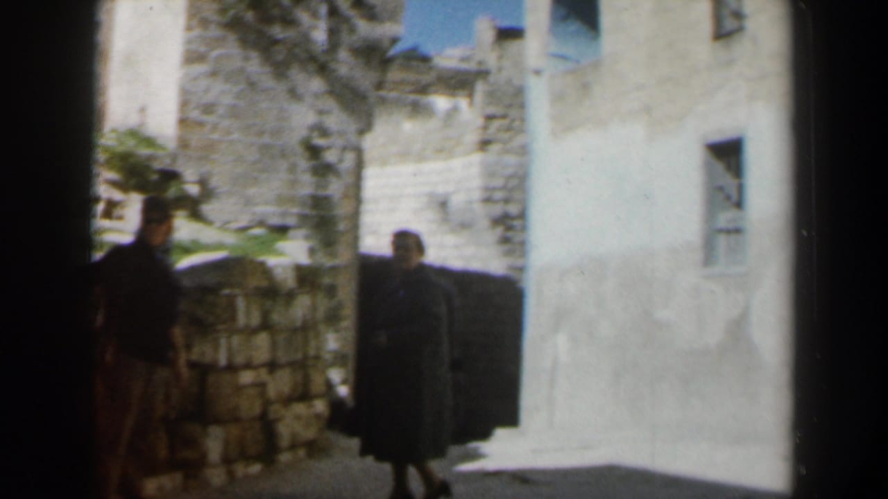 1960 Israel 8mm frame showing two adults partly in shadow beside stone walls and a pale plaster building.