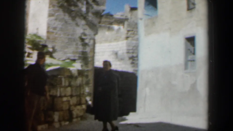 1960 Israel 8mm frame showing two adults partly in shadow beside stone walls and a pale plaster building.