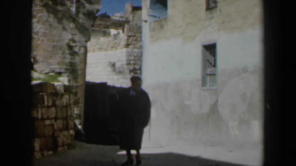 1960 Israel home-movie frame with an older woman in a dark coat near a narrow stone alley and pale wall.