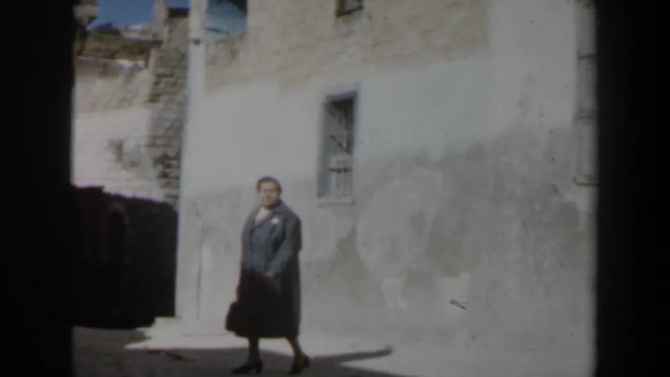 1960 Israel frame showing an older woman carrying a dark handbag through a narrow stone alley in strong sunlight.