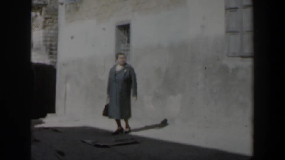 1960 Israel 8mm frame of an older woman in a coat holding a handbag beside a pale plaster wall and barred window.