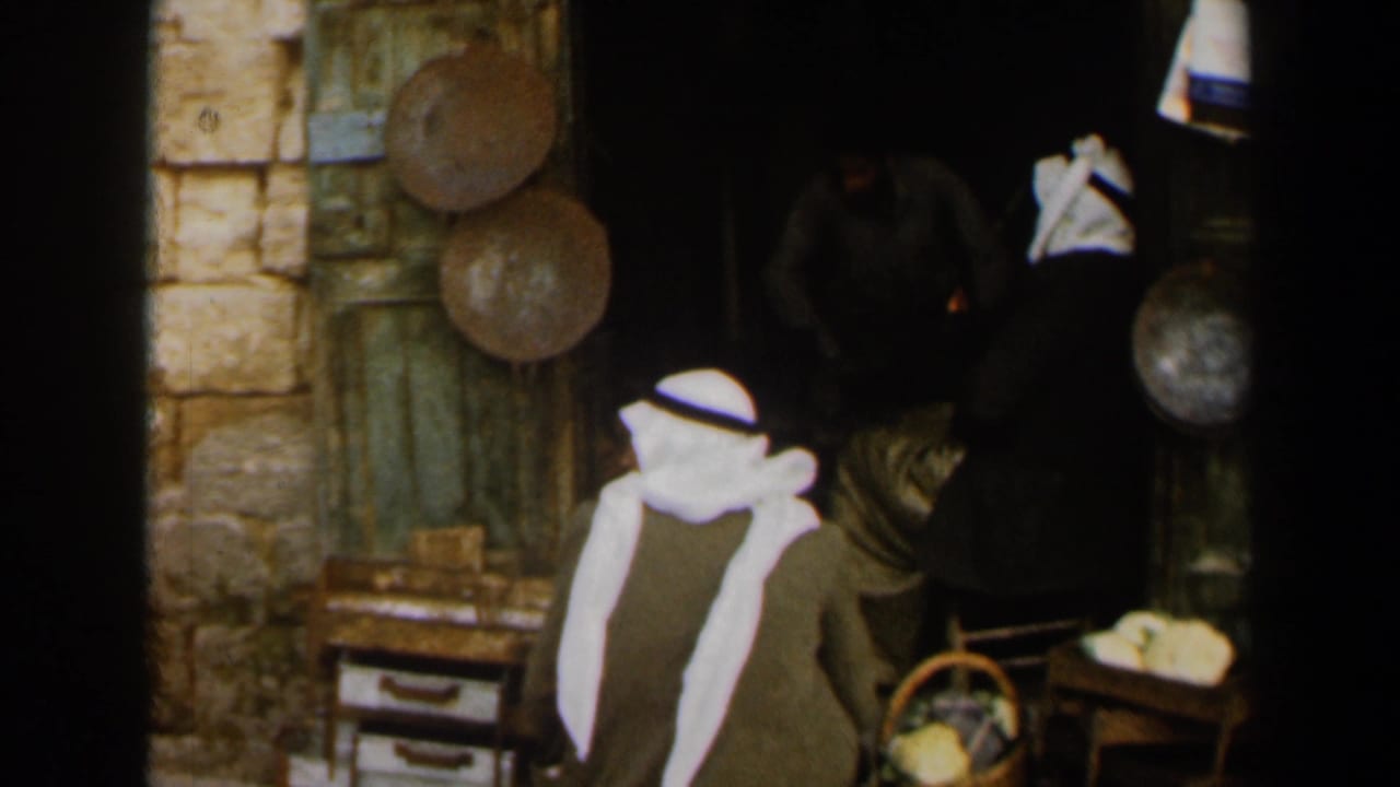 1960 Israel home-movie frame showing men in traditional attire near a weathered green shop entrance with baskets and metal pans.