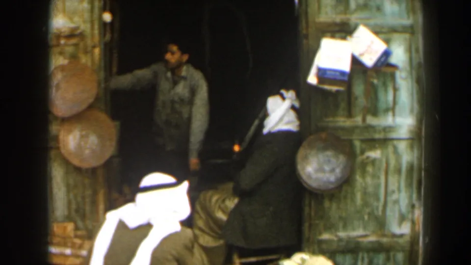 1960 Israel frame showing a man gesturing from a dark shop doorway near robed men and hanging wares on a green door.